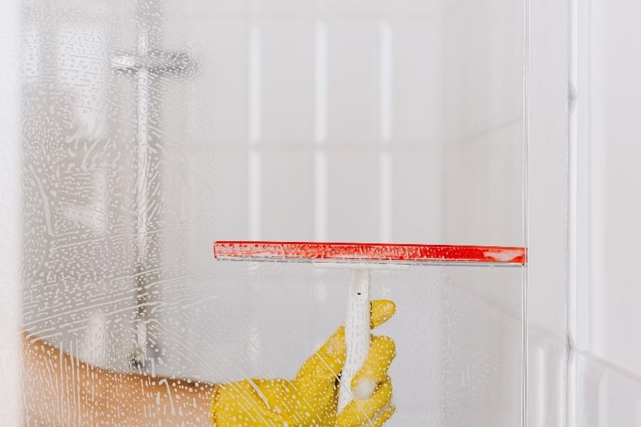 Yellow gloved hand using a red squeegee to wipe soapy water off a glass shower door in Austin TX