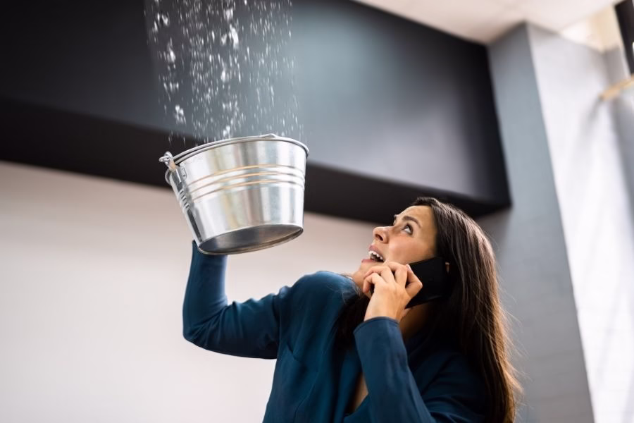 Homeowner on a phone call holding a bucket to catch water dripping from a ceiling leak in Austin TX