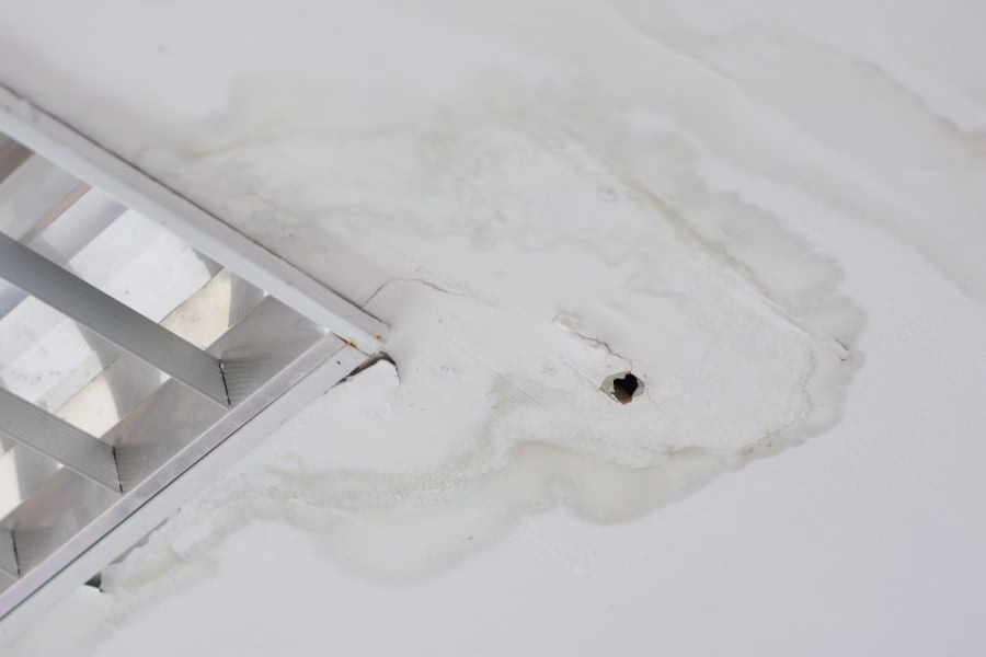 Water-stained ceiling drywall with visible drip pattern in a first-floor hallway, caused by an upstairs bathroom leak above