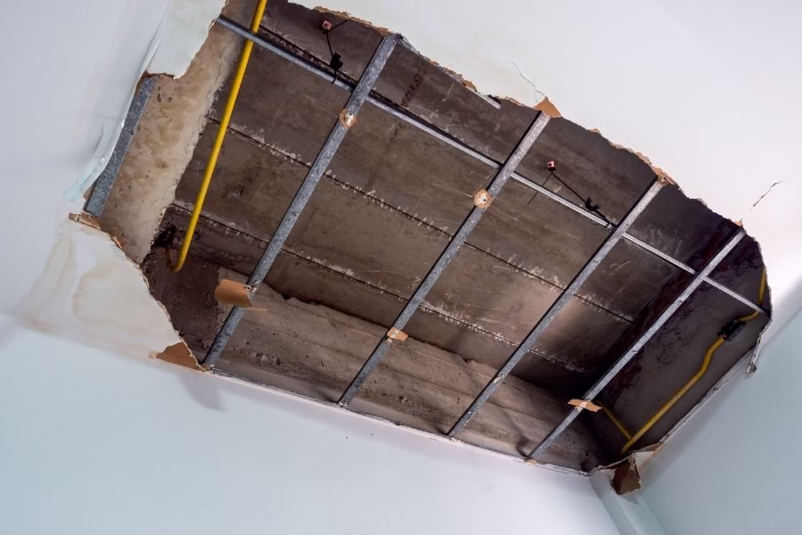 Technician on a step ladder cutting out a section of water-damaged ceiling drywall, wet insulation visible above the opening