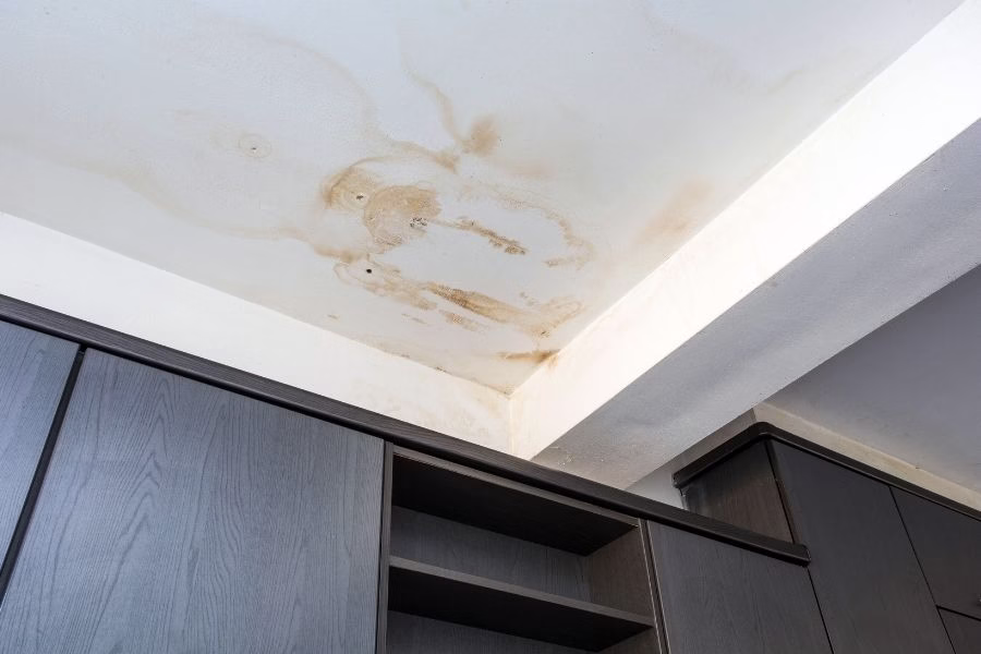 Brown water stain spreading across a white ceiling in a residential living room, with slight sagging visible near the center of the stain