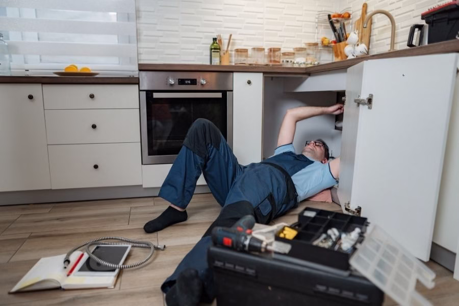 Technician with a flashlight inspecting moisture under kitchen cabinets, using a pin-type moisture meter on the cabinet base