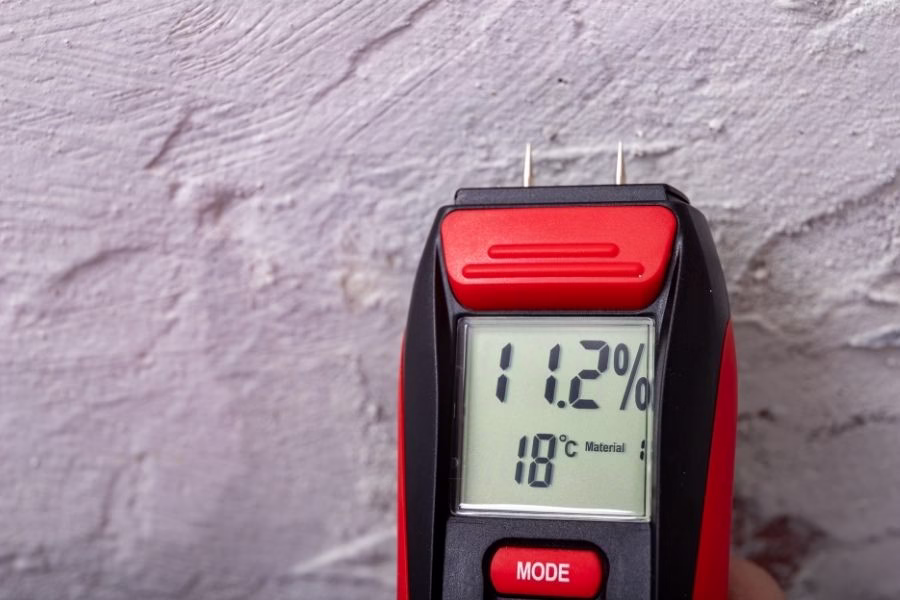 Technician using a non-invasive moisture meter on a painted wall in a hallway, with a digital reading showing elevated moisture levels