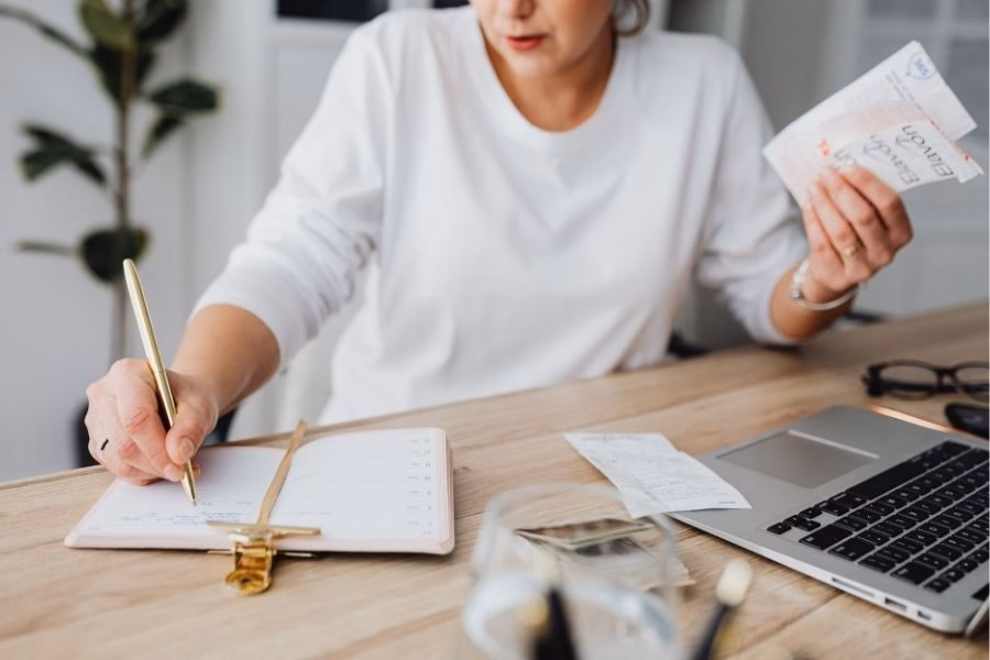 Woman writing a home inventory list at a desk while holding receipts next to a laptop